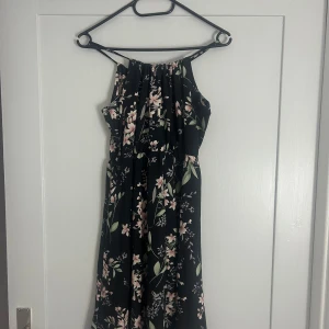 Svart medellångklänning med blommigt mönster - Säljer en svart medellångklänning med blommigt mönster i rosa och grönt. Klänningen har ärmlös design med halterneck och ett luftigt fall. Perfekt för sommarens alla tillfällen, med en skön och lätt känsla. Blommorna ger en romantisk vibe och klänningen är riktigt flowy.