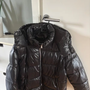 Svart glansig pufferjacka - Säljer en svart, glansig pufferjacka med stor huva från Lager 157. Jackan är slutsåld och väldigt populär till vintern. Den har en quiltad design, två fickor med dragkedja och justerbar nederkant. Perfekt för kalla dagar!! Pris går att diskuteras.