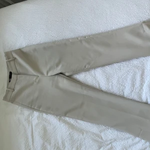 Beiga kostymbyxor - Snygga beiga kostymbyxor från Vila med klassisk rak passform och midja med hällor för bälte. Byxorna är tillverkade i ett mjukt och lätt material som ger en clean look. Perfekta för dig som gillar stilrena och enkla plagg.