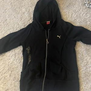 Svart hoodiejacka från Puma, strl L - Svart hoodiejacka från Puma med dragkedja och huva. Guldigt Puma-logo på bröstet och broderad Puma-text på sidan. Jackan har två fickor framtill och är i mjukt material, perfekt för chill eller träning. Köpt på second hand men tvättat efter köp, inte använd mer än 2 gånger.