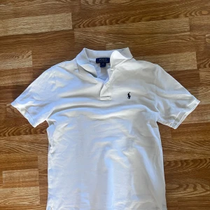 Vit pikétröja från Polo Ralph Lauren - Klassisk vit pikétröja från Polo Ralph Lauren med broderad logga på bröstet. Tröjan har korta ärmar, krage och är tillverkad i bomull. Perfekt för en clean och stilren look.