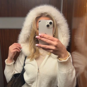 Vit stickad jacka med pälskantad luva - Supermysig vit stickad jacka med fluffig pälskantad luva och dragkedja framtill. Jackan har ribbade muddar och sitter snyggt med en figurnära passform. Perfekt för kalla dagar när du vill vara både varm och stylish.