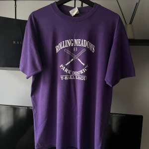 Lila t-shirt vintage  - Lila t-shirt från Jerzees med tryck 'Rolling Meadows Park District T-Ball League' och två korsade basebollträn på bröstet. Klassisk rund hals och korta ärmar. Materialet är en heavyweight blend som känns mjuk och slitstark. Perfekt för dig som gillar sportiga vintage-vibbar. Storleken är L men passa även mig som brukar bära storlek M. 