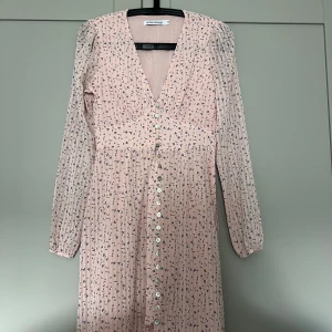 Ljusrosa blommig klänning från Adoore - Säljer en ljusrosa långklänning från Adoore med små blommigt mönster i blått och lila. Klänningen har v-ringning, långa ärmar och knappar hela vägen framtill. Perfekt för dig som gillar romantisk och feminin stil.