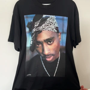 Svart 2Pac t-shirt med tryck - Svart t-shirt med stort färgtryck av 2Pac på framsidan. Modellen är loose fit med klassisk rund hals och korta ärmar. Materialet är mjuk bomull och trycket har coola streetvibes. Perfekt för dig som gillar hiphop och ikoniska motiv. Den är i ett bra skick. 
