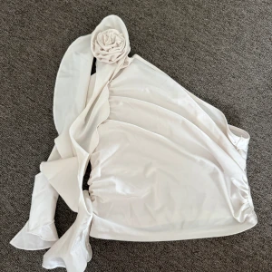 Vit oneshoulder topp med rosett - Säljer en superfin vit oneshoulder topp i glansigt material. Toppen har en stor dekorativ rosett på axeln och ett snyggt omlottliknande fall. Perfekt för dig som vill sticka ut med en unik och trendig look!