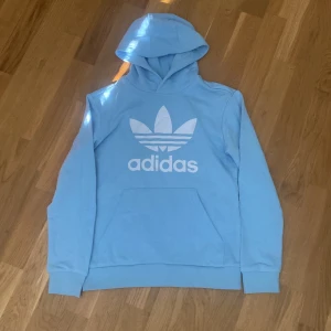 Ljusblå Adidas Trefoil hoodie - Säljer en ljusblå hoodie från Adidas med stor vit Trefoil-logga på bröstet. Tröjan har huva, magficka och ribbade muddar vid ärmslut och nederkant. Perfekt för chill dagar och streetstyle. Materialet är mjuk bomull som känns skönt mot huden.