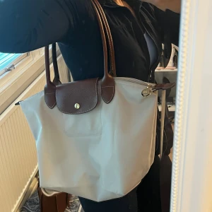 Beige axelväska från Longchamp -  Beige axelväska från Longchamp med bruna skinnhandtag och lock. Väskan har guldfärgad dragkedja med Longchamp-logga och är tillverkad i slitstark syntet med detaljer i skinn. Perfekt storlek får plats med dator och mycket mer. 