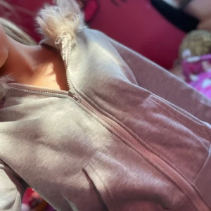 Grå hoodiejacka med fuskpälsluva - Mysig grå hoodiejacka med dragkedja och stor luva kantad med fluffig fuskpäls. Jackan har två framfickor och är tillverkad i mjuk bomullsmix. Perfekt för chill dagar eller när du vill ha en avslappnad stil.