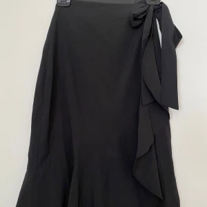 Svart omlottkjol från Gina Tricot - Svart långkjol från Gina Tricot i omlottmodell med snygg volangkant och knytband i midjan. Kjolen har en stilren look och är tillverkad i ett mjukt syntetmaterial som faller fint. 