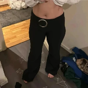 Svarta bootcut kostymbyxor med ringbälte - Svarta bootcut byxor med snyggt, stort ringbälte i silver. Byxorna har låg midja och vida ben, vilket ger en retro och trendig vibe. Perfekta för dig som vill sticka ut med en cool detalj i outfiten. Låna på mig som är 1.58😁