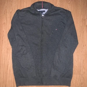 Mörkgrå kofta från Tommy Hilfiger - Snygg mörkgrå kofta från Tommy Hilfiger med dragkedja och ribbade muddar. Slim fit-modell med diskret logga på bröstet och ett randigt band i kragen i rött, vitt och marinblått. Perfekt för dig som gillar stilrena och klassiska plagg.