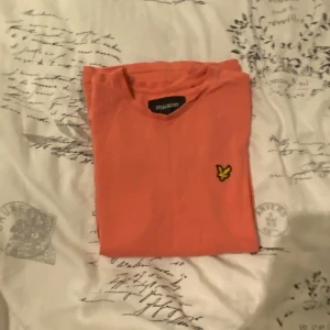 Korallfärgad t-shirt från Lyle & Scott - Snygg korallfärgad t-shirt från Lyle & Scott med klassisk gul fågel-logga på bröstet. Modellen är kortärmad och har rund hals. Materialet är mjuk bomull som känns skönt mot huden. Perfekt för dig som gillar stilrena och färgglada plagg.