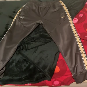 Nike svarta mjukisbyxor med guldiga detaljer - Snygga svarta mjukisbyxor från Nike med guldiga detaljer längs sidorna och Nike-logga på låret. Byxorna har elastisk midja och är tillverkade i ett glansigt syntetmaterial. Perfekta för chill eller träning, med en modern och sportig vibe.