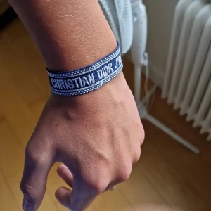 Blå Christian Dior J'adior armband - Snyggt blått armband från Christian Dior med texten 'J'adior' broderad i vitt. Armbandet har en justerbar knytning och är gjort i ett mjukt tygmaterial. Perfekt accessoar för att lyfta din outfit med lite lyxig vibe. No size, passar alla. Storleken kan justera genom att dra i snörena 🍾😄