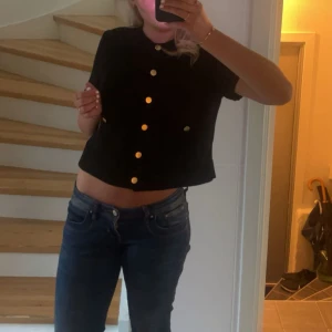 Svart croppad kofta med guldfärgade knappar - Svart stickad kofta från H&M med korta ärmar och snygga guldfärgade knappar framtill. Ribbad kant vid hals och ärmslut, samt två fickor framtill. Perfekt för dig som gillar stilrena detaljer och vill ha en modern look. Perfekt nu till hösten, men funkar såklart året om☺️tröjan är i bra skick och i storlek S!