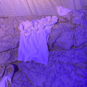 Vit oneshoulder topp med volang Kappahl - Snygg vit oneshoulder topp från Kappahl med volangdetalj längs halsringningen. Toppen är ärmlös och har en ribbad struktur som ger en cool touch. Perfekt för dig som gillar att sticka ut med en unik och trendig stil.