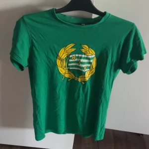 Hammarby t-shirt  - Grön t-shirt från HIF med klubbens emblem tryckt i gult och vitt på bröstet. Klassisk rund hals och korta ärmar. Tillverkad i mjuk bomull, perfekt för dig som vill visa ditt stöd för laget. Snygg och enkel design med sportig vibe.