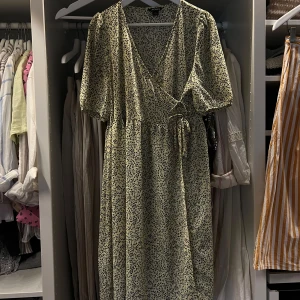 Blommig omlottklänning från Monki - Supersöt långklänning från Monki i ljusbeige med svart blommigt mönster. Omlottmodell med v-ringning, korta puffärmar och knyt i midjan på inne i klänningen och utanpå. Perfekt för dig som gillar flowy vibes och vill ha en klänning som sticker ut lite extra och ger en superfint siluett. 