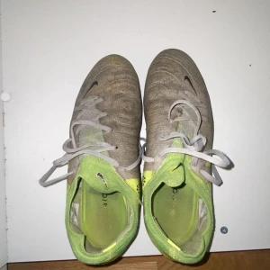 Nike fotbollsskor i gul och grå - Säljer ett par Nike fotbollsskor med snygg design i neon-gult och grått. Skorna har snörning och en svart Nike-logga på sidan. Ovandelen är tillverkad i syntetmaterial och har en rund tå samt platt sula med dobbar för gräsplan. Perfekt för dig som vill sticka ut på planen.