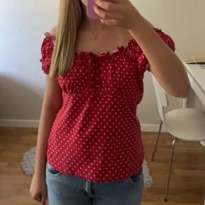 Röd prickig offshoulder blus - Supersöt röd blus med vita prickar och offshoulder-modell. Toppen har korta puffärmar, volangdetaljer och en liten knytning framtill. Tillverkad i mjuk viskos med lite stretch för en bekväm passform. Perfekt för dig som gillar en lekfull och trendig stil.