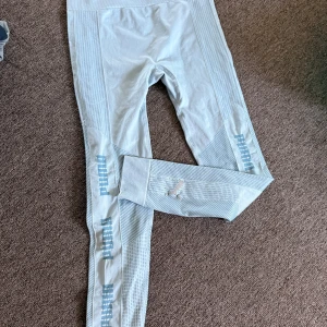 Ljusblå leggings från Puma - Snygga ljusblå leggings från Puma med randiga detaljer längs benen och logga tryckt i blått. Materialet är mjukt och stretchigt, perfekt för träning eller chill. De har en tight passform och hög midja som sitter skönt. Puma-loggan syns även på vaden för extra sportig vibe.