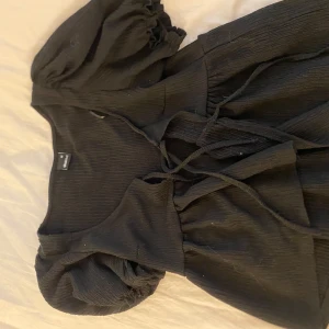 Svart omlottklänning med puffärm - Säljer en svart omlottblus från Gina tricot med puffiga kortärmar och snörning i midjan. Blusen har en lätt ribbad struktur och ger en snygg siluett. Perfekt för dig som gillar stilrena och trendiga plagg.