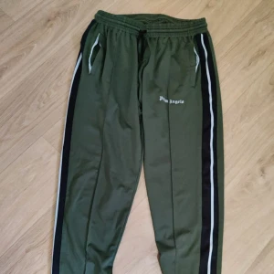 Palm Angels gröna mjukisbyxor - Snygga gröna mjukisbyxor från Palm Angels med svarta och vita ränder längs sidorna. Byxorna har resår och snörning i midjan samt fickor med vit kant. Perfekta för en chill och sportig look.