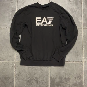 Svart EA7 Emporio Armani sweatshirt - Svart långärmad sweatshirt från EA7 Emporio Armani med stor vit logotyp på bröstet. Tröjan har rund hals och ribbade muddar vid ärmslut och nederkant. Tillverkad i mjuk bomull som är skön mot huden. Perfekt för dig som gillar stilrena och sportiga märkeskläder. Storlek M men passar även S! Hör av dig vid frågor eller funderingar!