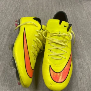 Vapor 10 Nike Mercurial gula fotbollsskor - Säljer ett par Nike Mercurial fotbollsskor i en riktigt skarp gul färg med svarta och orange detaljer. Skorna har snörning, låg profil och platt sula med dobbar för gräsplan. Insidan är orange och hälen har Mercurial-tryck. Perfekt för dig som vill sticka ut på planen. Använd typ 4 ggr