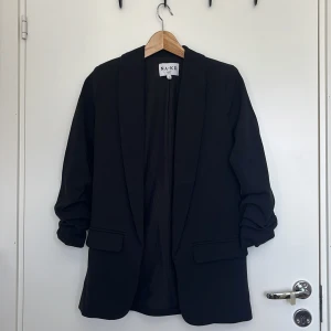 Svart blazer från Nelly strl 32 - Svart öppen blazer från Nelly i storlek 32. Uppsrunchade armar, lite kortare armar, tre kvarts längd ish. Aldrig använd. 