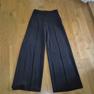 Svarta wide kostymbyxor från H&M - Snygga svarta kostymbyxor från H&M med breda ben och hög midja. Byxorna har hällor för bälte, dold dragkedja och hake framtill. Perfekta för en clean och stilren look. Materialet är mjukt och följsamt, vilket gör dem riktigt bekväma. Har fickor ifram.