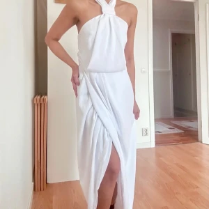 Vit långklänning från Shona Joy - Elegant vit långklänning från Shona Joy med halterneck och omlottdesign. Klänningen har en slits framtill och stängs med dragkedja i sidan. Perfekt för sommarens alla tillfällen där du vill känna dig extra snygg.