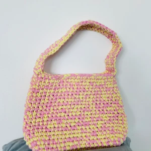 Virkad axelväska i rosa och gult - Superfin stickad axelväska i pastellrosa och gul. Väskan har en chunky, handgjord känsla med bred axelrem och mjuk struktur. Perfekt för dig som vill ha en färgglad accessoar som sticker ut.