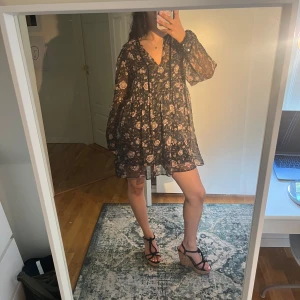 Svart blommig kortklänning  - Supersöt svart kortklänning med rosa och beige blommor, v-ringad och med snörning vid halsen. Klänningen har långa, transparenta ärmar och ett flowigt, loose fit. Den är perfekt för tex sommarkvällar!😘