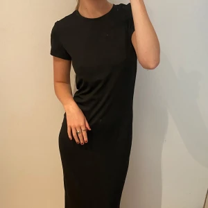 Svart långklänning med kort ärm - En stilren svart långklänning med rund hals och korta ärmar. Klänningen har en rak och slim passform som ger en snygg siluett. Perfekt för dig som gillar minimalistisk och enkel design. Materialet känns mjukt och bekvämt mot huden.