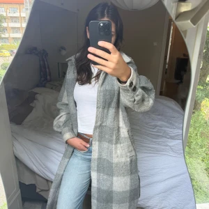 Grå rutig lång overshirt - Säljer en lång och oversized grå overshirt med rutigt mönster. Jackan har stora knappar vid ärmsluten, klassisk krage och två fickor framtill. Perfekt att slänga över en hoodie eller t-shirt för en chill look.