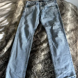 Ljusblå jeans från Scotch & Soda - Säljer ett par ljusblå jeans från Scotch & Soda Amsterdam Blauw, tillverkade i ekologisk bomull. Klassisk femficksmodell med raka ben och normal passform. Snygga detaljer med läderpatch bak i midjan och subtila slitningar vid fickorna.