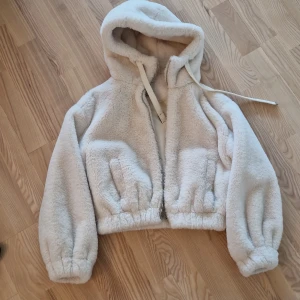 Vit teddyjacka med huva och dragkedja - Supermysig vit teddyjacka från Bershka med stor huva och dragkedja framtill. Jackan har elastiska muddar vid ärmslut och midja, samt två fickor på sidorna. Perfekt för kalla dagar när du vill vara både varm och snygg. Fodrad insida för extra komfort.