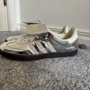 Adidas Samba Wales Bonner  - Unika Adidas Samba Wales Bonner sneakers i spegelblankt silver med krämfärgade detaljer och stickade ränder. Skorna har en brun sula, snörning och extra stor plös med logga. Materialmix av syntet och textil ger en riktigt futuristisk vibe.
