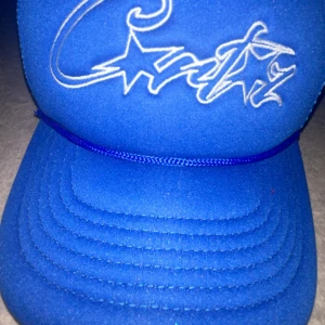 Blå Corteiz truckerkeps med broderi - Säljer en blå truckerkeps från Corteiz med vit broderad logga och stjärnor framtill. Kepsen har mesh på sidorna och bak, samt ett snyggt rep över skärmen. Insidan har Corteiz-tryck på banden och en gul etikett. Perfekt för dig som vill sticka ut.