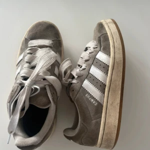 Adidas Campus - Snygga Adidas Campus sneakers i grå mocka med vita klassiska ränder och vit sula. Skorna har rund tå, platt sula och snörning. Perfekta för dig som gillar retrostil och streetwear. Storlek 36,5 (EU).
