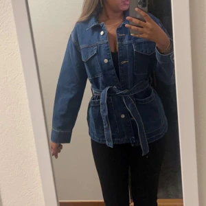 Mörkblå jeansjacka med bälte - Snygg mörkblå jeansjacka med knappar framtill och matchande bälte i midjan. Jackan har två stora bröstfickor och klassisk krage. Perfekt för dig som gillar en avslappnad men trendig look.