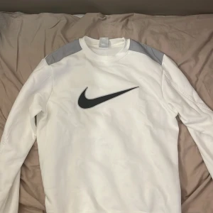 Vit Nike sweatshirt med grå axelpartier - Nike sweatshirt i vitt med stora grå axelpartier och en svart Swoosh-logga på bröstet. Tröjan har rund hals, långa ärmar och är tillverkad i mjuk bomullsmix. Perfekt för en sportig och clean look. Helt ny och aldrig använd. 