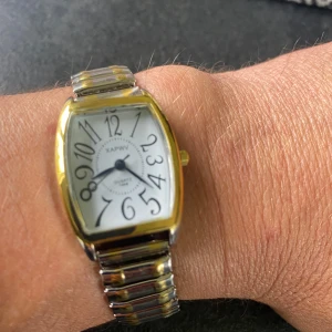 Xapnw klocka i guld och silver - Snygg klocka från Xapnw med rektangulärt boett i guld och silver. Urtavlan är vit med svarta, lekfullt snirkliga siffror och blå visare. Armbandet är elastiskt i silver och guld, vilket gör den enkel att ta på och av. Perfekt accessoar för dig som gillar unik design.