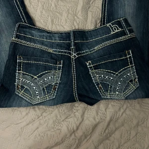 Hydraulic jeans med broderade fickor - Mörkblå jeans från Hydraulic med kontrastsömmar och snygga broderade bakfickor med silvriga detaljer. Klassisk femficksmodell i jeansmaterial, låg midjade.Lite trasigt gylf men mycket lätt att fixa.Perfekta för dig som gillar statement-jeans med coola detaljer.