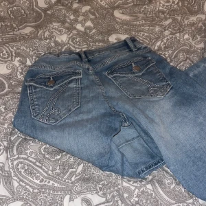 Blå jeans med detaljerade bakfickor - Säljer ett par ljusblå bootcut jeans från lindex med snygga kontrastsömmar och unika bakfickor med lock och knapp. Jeansen har klassisk femficksdesign, bälteshällor och dragkedja framtill. Perfekta för dig som gillar jeans med lite extra detaljer.