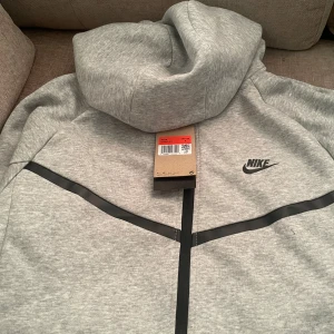Grå Nike hoodiejacka med dragkedja S - Snygg grå hoodiejacka från Nike med svart logga på bröstet och svarta detaljer längs dragkedjan. Jackan har huva och hel dragkedja framtill, perfekt för en sportig och chill stil. Tillverkad i mjuk bomullsblandning för extra komfort.
