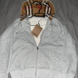Grå Burberry hoodie med dragkedja - Pris kan diskuteras.Snygg grå hoodie från Burberry med klassiskt rutigt mönster i huvan. Tröjan har dragkedja framtill, snörning vid huvan och broderad Burberry-logga på bröstet. Tillverkad i mjuk bomull och har praktiska fickor framtill. Perfekt för chill dagar.Står att det är L men sitter mer som S/M