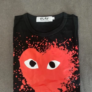 Svart t-shirt med rött hjärta från Comme des Garçons PLAY - Svart t-shirt från Comme des Garçons PLAY med stort rött hjärta och ögon framtill. Häftigt stänkmönster runt hjärtat som ger en unik look. Klassisk rund halsringning och korta ärmar. Perfekt statement-plagg för dig som gillar streetstyle.